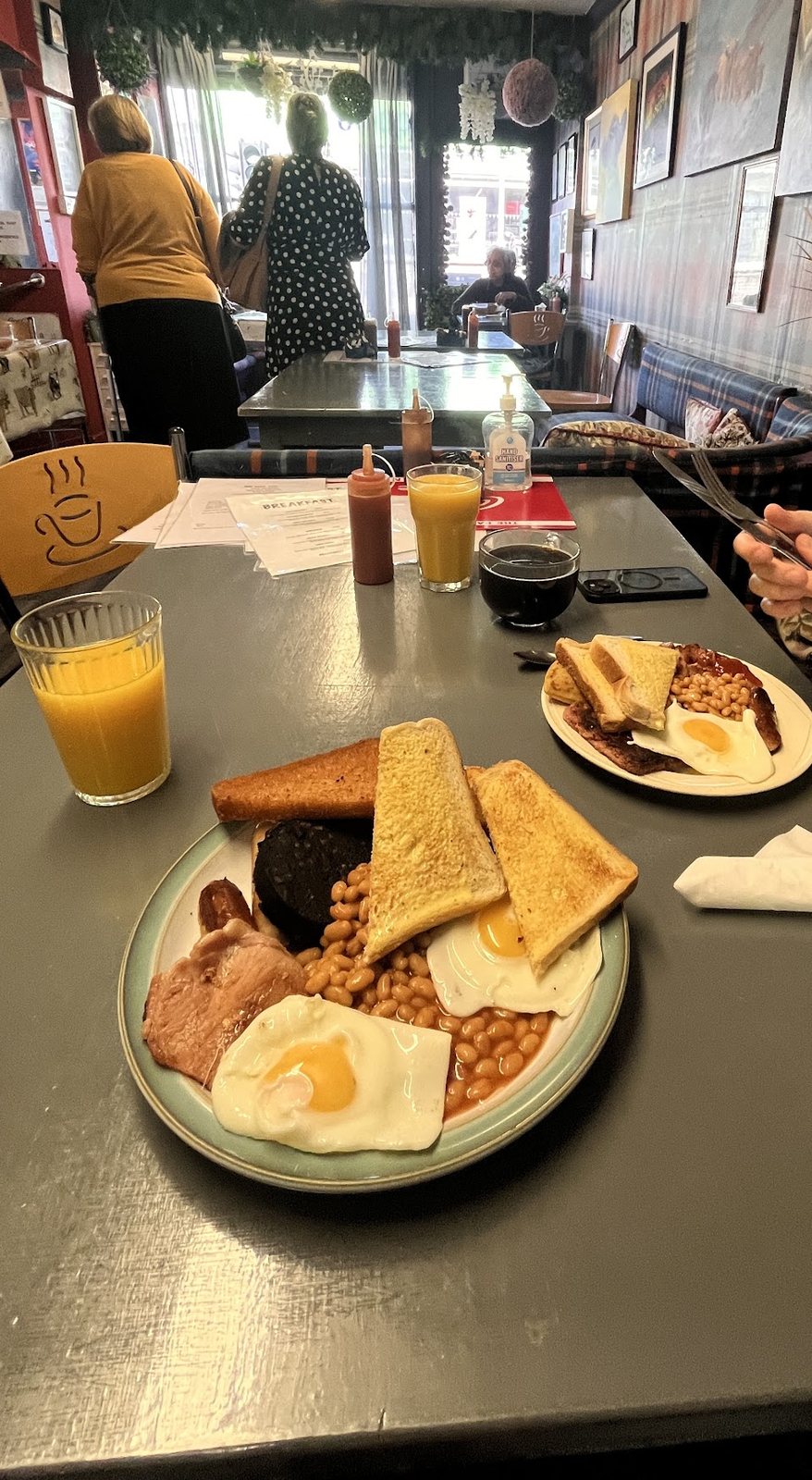 A full breakfast with eggs, beans, black pudding and toast, and an orange juice, on a grey table in the tartan-walled café.
